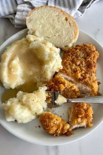 A plate of ranch chicken, mashed potatoes and gravy, with a slice of french bread with butter