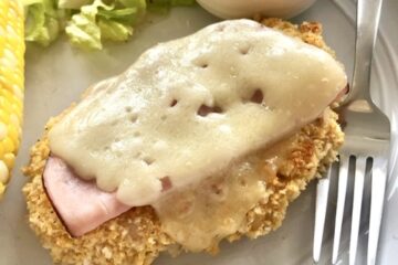 oven baked breaded chicken topped with deli sliced ham and swiss cheese served on a plate with honey mustard dipping sauce, a side salad and corn on the cob