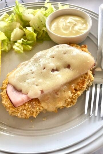 oven baked breaded chicken topped with deli sliced ham and swiss cheese served on a plate with honey mustard dipping sauce, a side salad and corn on the cob