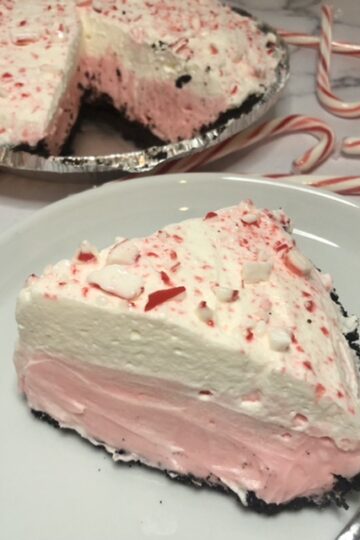 slice of no bake peppermint pie with Oreo chocolate crust and peppermint pie filling and topped with whipped cream and crushed candy canes, and the pie in the background with the slice taken out of it