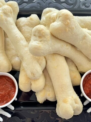 Bones breadsticks on a black serving platter with plastic skeleton hands as decoration with marinara sauce in a small dish for dipping sauce that looks like blood for Halloween