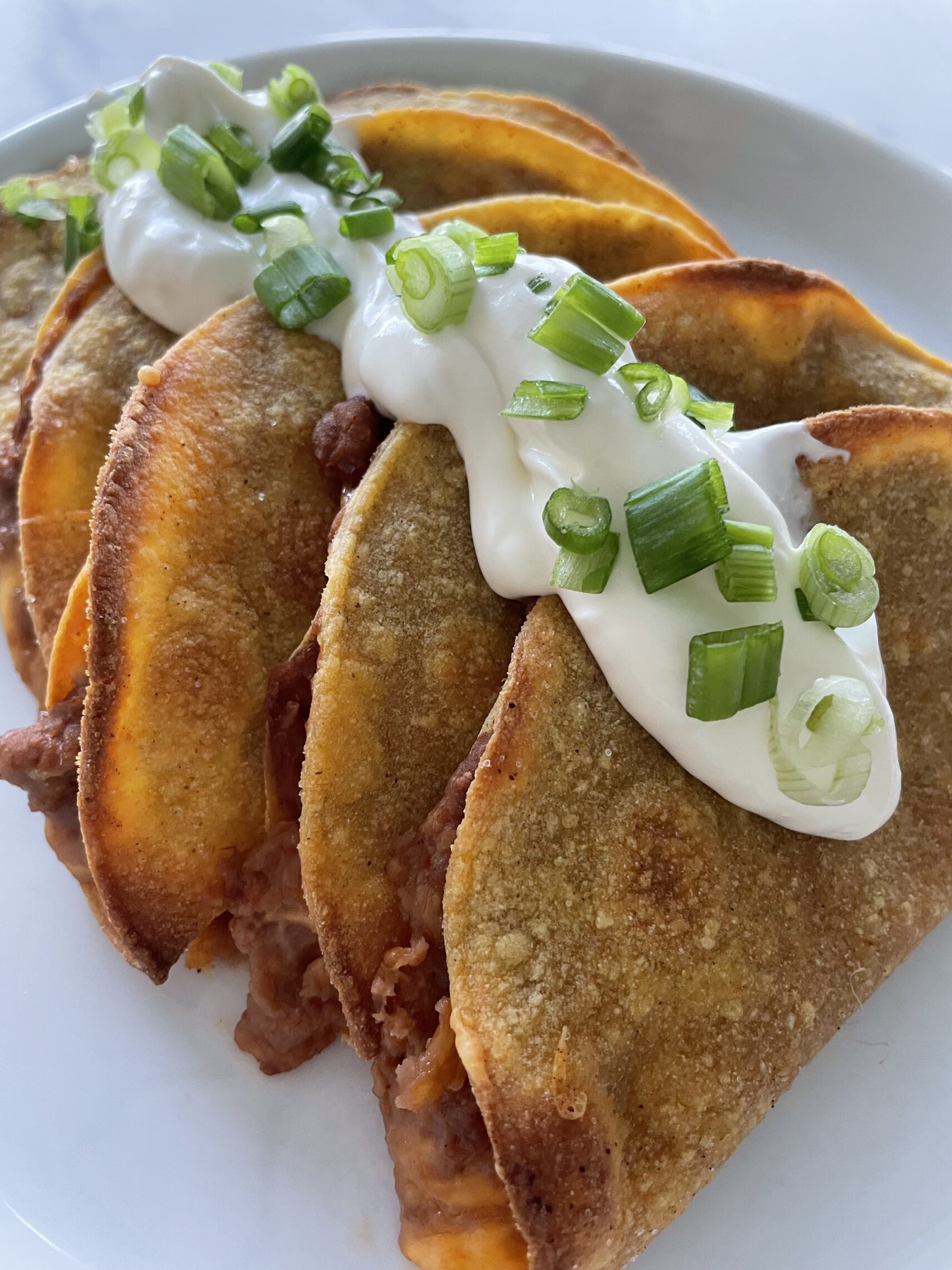 A plate full of crisp bean tacos with sour cream and chopped green onions on top.